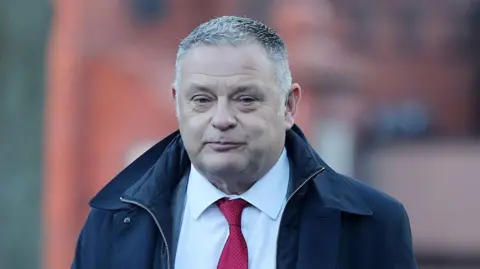 Mike Amesbury, with grey hair and clean shaven, is wearing a dark blue coat, white shirt and red tie.