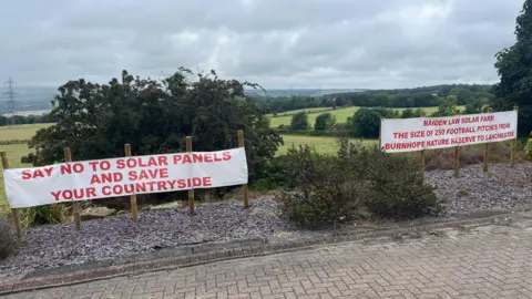 Two white banners with red letters attached to a fence on the edge of fields. One reads: "Say no to solar panels and save your countryside" and the other says: "Maiden law solar farm the size of 250 football pitches from Burnhope nature reserve to Lanchester".