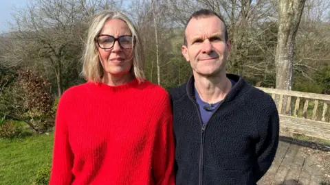 Juliette Parkin/BBC Tracey and James Parfitt pictured at her home in Lamberhurst, Kent. Tracey is wearing a red jumper and is looking at the camera. She is wearing a red jumper and has blonde hair. James is wearing a blue t-shirt and black fleece. He has brown hair.