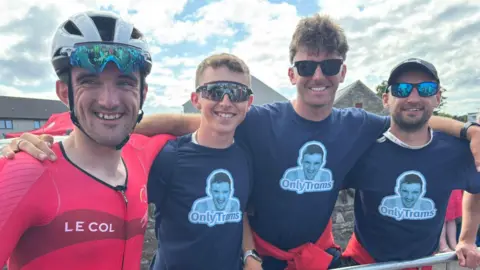 Manx Radio Sport Tyler Hannay wears a red vest and a white helmet, which has sunglasses attached. He stand with his arm around three men who wear navy blue t-shirts with Hannay's face on. 