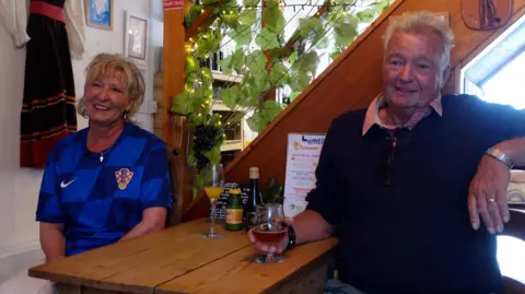 Suzi sits beside Ed’s father Andy at a wooden table indoors, with Suzi wearing a blue Croatia football shirt and Andy holding a glass, with plants, lights and decorations around them.