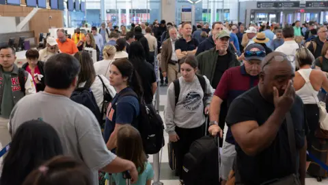 Lines of passengers are shown inside an airport stretched across the check-in area. People look tired and frustrated. 