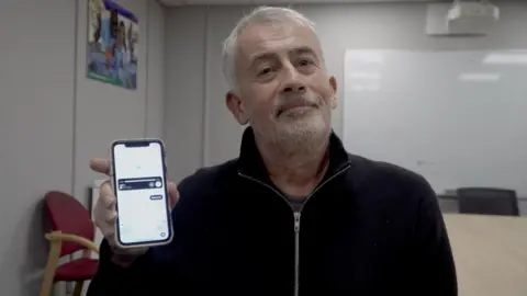 A man with short, white hair and a white beard smiling at the camera. He is wearing a black jacket and holding a phone, showing the screen to the camera.