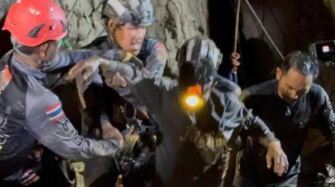 Two rescuers in dark blue diving gear, with one wearing a black helmet and the other a red one, hold the right arm of the rescued man, who is dark muddy clothes and has a headlight on. His left arm is being held by another rescuer to his left as they all emerge from the cave in darkness
