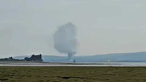 Victoria Mcmurtha A large cloud of smoke rises above the scene. Ancient ruins can be seen on the left by an expanse of water and grass in the foreground. Hills form the backdrop.