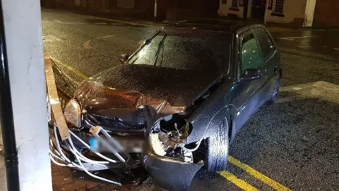 San Tse  A black car with a crumpled front that has smashed into the restaurant. The street railings appear to be crumpled under the bonnet.