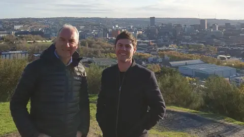 Sheffield Council Two men in dark coats stand smiling at the top of a grass bank with a city stretching out below them