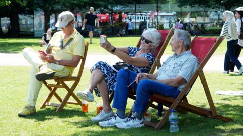Three people in deckchairs in a sunny urban park - two are posing for a selfie, the third is looking at a phone