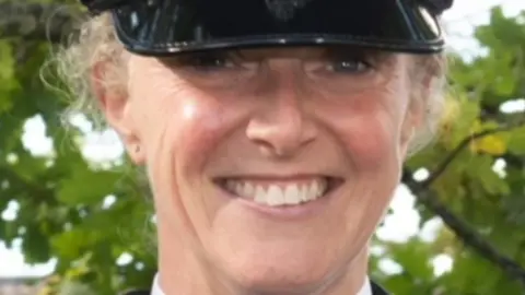 North Yorkshire Police A police officer in her forties, smiling directly at the camera. She is wearing a police cap which comes down to just above her eyes. She is pictured against the backdrop of some trees.