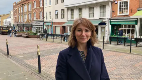A picture of Lisa Spivey in a navy blue jacket smiling at the camera. She is on a street with many shops behind her.
