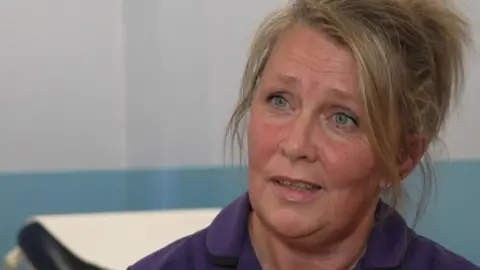 A nurse with blonde hair tied up and wearing a purple top. She is talking in a hospital consulting room.