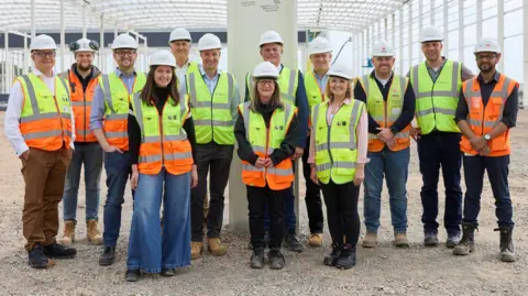 A large group of people standing in the middle of a construction site, all wearing white hard hats, and high-viz jackets, smiling and looking straight at the camera. A white steel post is behind them and scaffolding. 