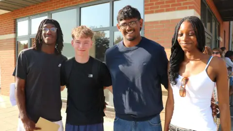 A group of four pupils standing outside a school building in the sunshine, smiling to camera. 