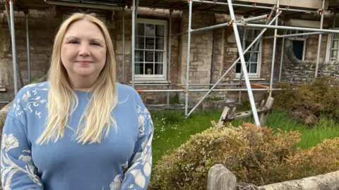 Melanie is standing outside her home on Vicars' Close. Melanie has blonde hair and is wearing a blue jumper with white flowers. Scaffolding surrounds the building behind her. 