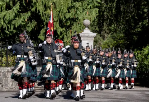 PA Media Soldiers from the Royal Regiment of Scotland march in formation during a ceremonial parade, carrying rifles and wearing dark dress uniforms, tartan kilts and white spats. Union flag is carried near the front of the column as the troops advance along a tree-lined route past a stone entrance.