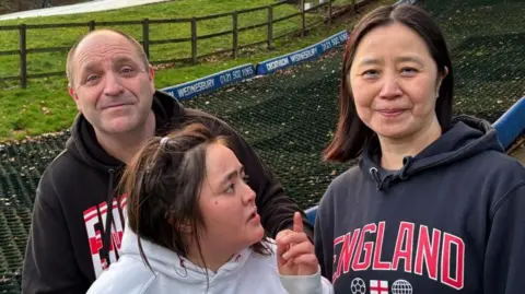 There are three people called Simon, Mandy and their daughter Emma. The woman on the right (Mandy) is wearing a dark grey hoodie with the word England written on it with big red letters. She has dark brown shoulder-length hair. In the middle, there is a young woman (Emma) looking up towards her mum. She is wearing a white jumper and has long dark brown hair. The man on the left (Simon) is wearing a black jumper with red and white lettering. The letters EN are visual. He has a grey hair in a buzz-cut hairstyle and is balding.