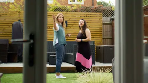 Two women are standing and talking in a garden area, seen through a glass door. The garden has a wooden deck with dark brown furniture, a wooden fence with lattice on top, and green grass in the background. There is a plant with spiky leaves near the bottom of the picture, and a brick building partially in the background.