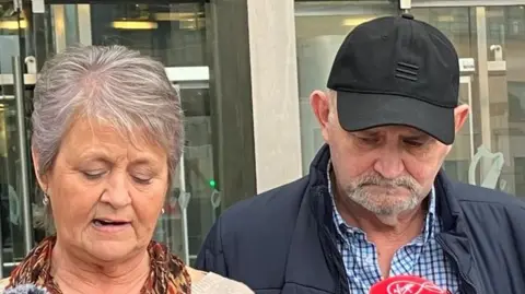 A woman with short grey and blonde hair is speaking into a mic as she wears a brown patterned scarf and a neutral coloured sweatshirt.  A man stands next to her wearing a black cap, and has grey facial hair. He is also wearing a blue checked shirt and black coat.