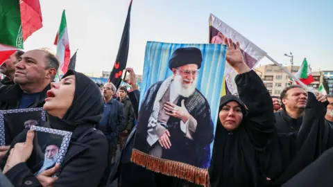 Protesters in Iran hold up flags and banners with Khamenei's picture on them. A woman in a black burqa hold her hand in the air and aother lady isclutching an image of Khamenei to her chest