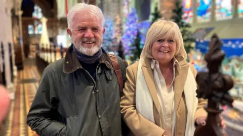 Tony and Aileen Moore, wearing winter overcoats, stand in the aile of st Columbs Cathedral, as the lights of the church's christmas festival twinkle in the background.