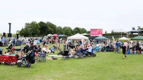 The Jockey Club Lots of people sit in groups on chairs and under gazebos on a grassy area at a racecourse. 