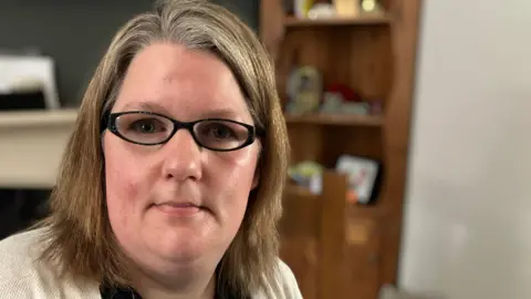 Samantha O'Sullivan to the left wearing glasses with shoulder length hair. There is a dark wooden cabinet in the background.