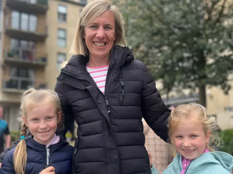 A woman with blonde hair in the centre of the image, smiling at the camera, with her two young daughter either side of her 