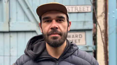 Daniel is standing outside in front of a blue fence. He is wearing a black cap and coat. He has a black beard. 