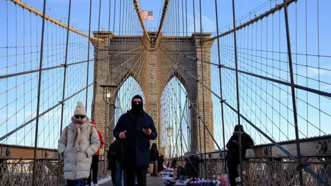 Pedestrians walk across the Brooklyn Bridge in New York City. One woman wears a beige puffer coat, a wooly scarf, sunglasses and a wooly hat with her hands in her pocket. Next to her, a man is dressed in a black balaclava, long black puffer coat and trousers. Around them, the suspension cables of the bridge connect to a pylon with the United States flag flying atop it. The sky is clear and blue.