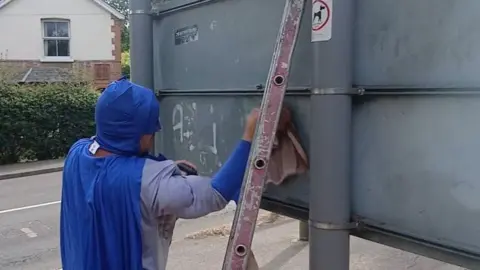 Liam Keen A man dressed as Batman cleans graffiti from a street sign.