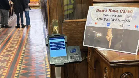 An electronic tablet is fixed to a stand at the inside door of a church. To the left, legs and feet can be seen standing up a tiled aisle. To the right, on top of wooden unit there is a sign that says: "Don't have cash? Please use our contactless card terminal." 