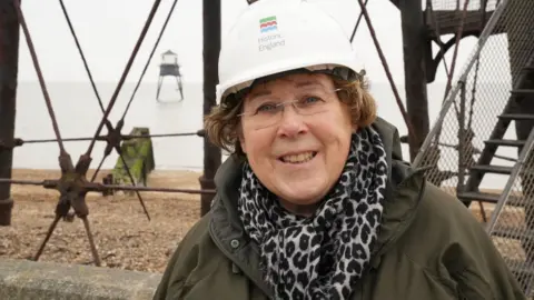 John Fairhall/BBC Trudi Hughes from Historic England, standing next to one of the Dovercourt lighthouses, with the second lighthouse pictured over her shoulder out at sea. Trudi is wearing a white hard hat with Historic England branding on the front. She is wearing glasses and smiling at the camera. Trudi is wearing a dark green coat with a leopard-print scarf