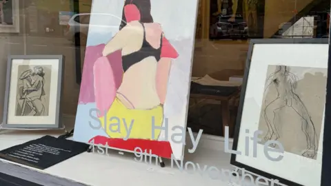 Val Harris Picture of the gallery window display, one large canvas in centre of a woman's bare back, a framed line drawing of a naked man (right) and a framed image of a hunched over naked woman (left). 