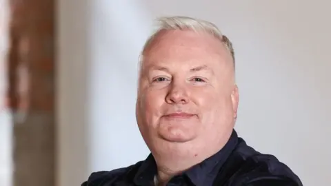 Stephen Nolan filmed in a room with red brick and white walls. He has short grey hair and a black shirt. He is standing with folded arms