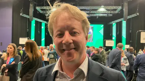 Shariqua Ahmed/BBC Paul, a man, with blonde hair, wearing a grey suit, smiling for camera.