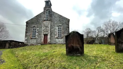 Wamphray Church is sitting on a hill in a countryside setting with headstones around it.