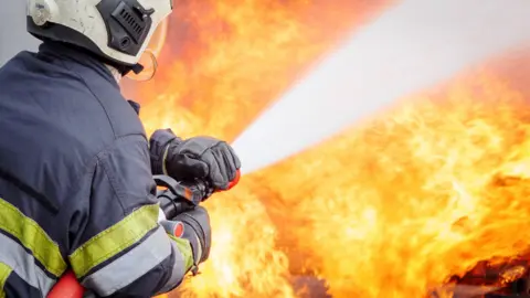 A firefighter wearing protective clothing holding a hose to battle flames