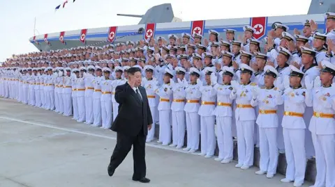 Rows of soldiers in white uniforms and white caps cheer as Kim Jong Un walk in front of them, wearing a black suit and raising his hand. Behind them is a massive grey ship.
