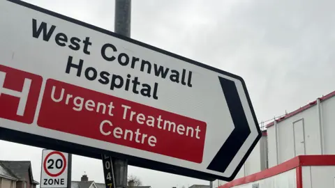 A sign which reads West Cornwall Hospital Urgent Treatment Centre. Behind it are portacabins and a 20mph speed sign. 