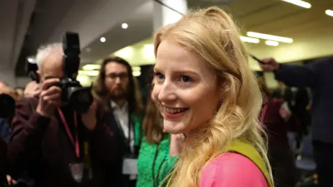EPA/Shutterstock Green Party candidate Hannah Spencer reacts after the vote count for the Gorton and Denton by-election in Manchester. She has long blone hair and is wearing pink and green
