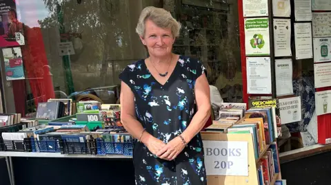 A woman in a black dress with a blue pattern standing in front of a community centre with her arms folded in front of her.