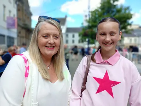 A middle aged woman with long blond hair, wearing a white top, stands with a teenage girl with brown hair parted on the middle who is wearing a light pink top with a star on it