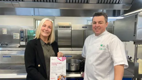 The Guernsey Institute Jane Fears is wearing a black jacket with a gold and black blouse. She has shoulder-length straight blonde hair. She is standing with Rivelino Rodrigues, who is a chef lecturer at TGI College. He is wearing his chef whites and has short dark hair. Both people are standing in a kitchen with silver units.