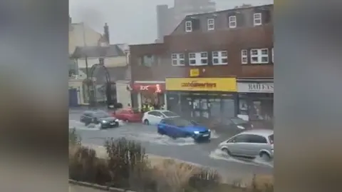 Rudd1979/X Cars driving in floodwater in front of shops on Dunstable High Street.