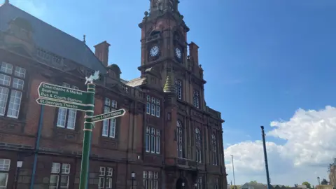 Owen Sennitt/BBC A large civic building with red bricks, a clock and tower can be seen with blue skies in the background
