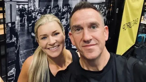 A woman with long blonde hair, and a man with short dark hair both look directly into the camera. There is a large black and white image of a group of people behind them