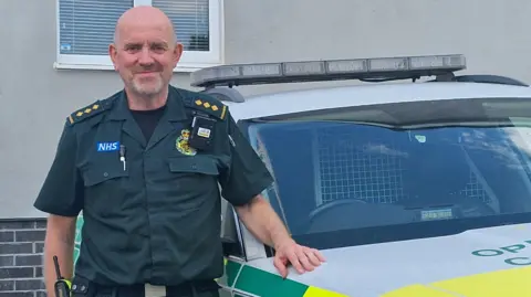 The image shows a paramedic stood in a the usual NHS ambulance service dark green uniform standing next to an emergency vehicle. He is looking straight at the camera and smiling while resting his hand on the car. 