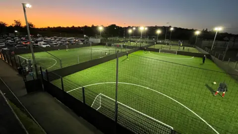 A series of five-a-side football pitches at Imperial Sports Centre in Bristol are seen from a high vantage point. The floodlights on and the distant sky has a tinge of orange as the sun goes down. There are a handful of players kicking footballs around on some of the pitches, which are covered in green astroturf.