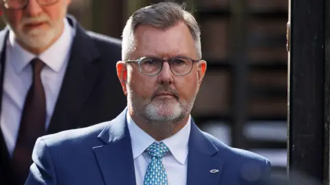 PA Media Sir Jeffrey Donaldson - a man with grey hair wearing small grey, circular glasses and a beard. He is wearing a navy suit, a white-collared shirt and a blue and white patterned tie. On the lapel is a small, ichthys symbol pin.