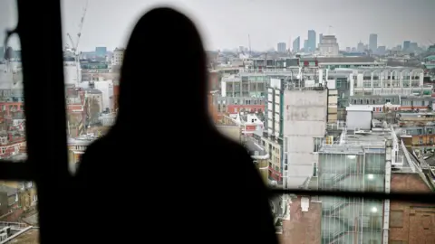 BBC Beth, pictured in a blurred silhouette against a high window, looking out onto tall buildings stretching into the distance on an overcast day